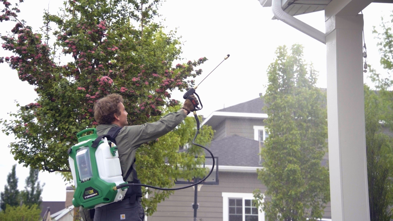 Professional pest control service technician spraying exterior home treatment with backpack sprayer near gutters and roofline, showing residential pest management and preventive maintenance in action