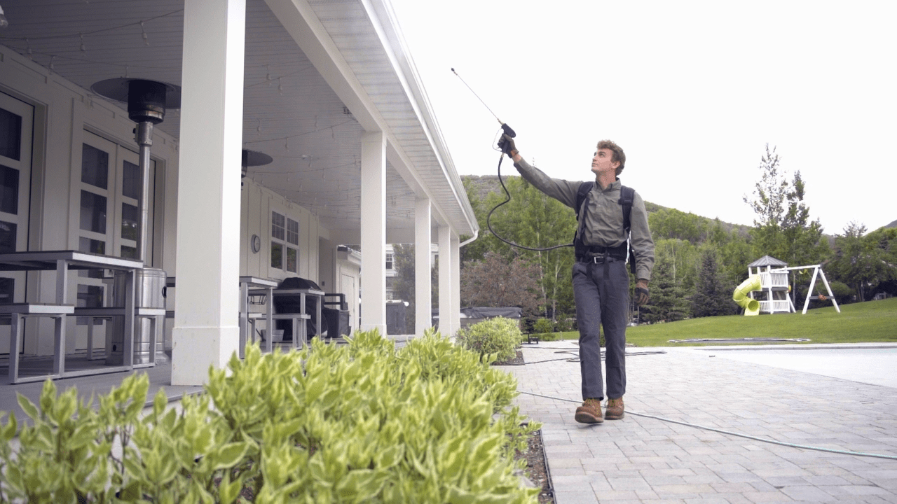 A pest control technician using a long-reach wand to treat the eaves of a modern white Utah residential home.