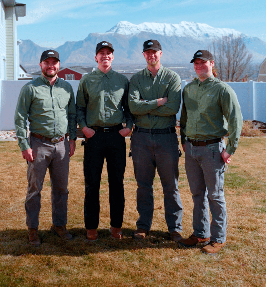 Four Nature's Balance Pest Control team members in matching green button-up shirts and black branded caps smile for a group photo outdoors, with a snow-capped Utah mountain and blue sky in the background.