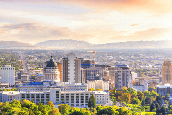 Aerial view of Salt Lake City, Utah during sunset. The foreground shows the Utah State Capitol building with its distinctive dome, surrounded by autumn trees with gold and green foliage. Downtown skyscrapers rise beyond the Capitol. In the background, the Wasatch mountain range stretches across the horizon under a colorful sky with pink and orange clouds.