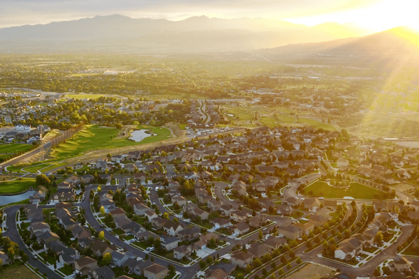 Aerial view of Cedar Hills, Utah at sunset or sunrise. The image shows a planned suburban neighborhood with curved streets and residential homes arranged in a circular pattern. A golf course with green fairways and small ponds is visible near the housing development. In the background, mountains rise against a golden-lit sky with scattered clouds, while the sun casts a warm glow across the valley. The perspective appears to be from a drone or elevated position overlooking the community with both developed residential areas and open spaces visible.